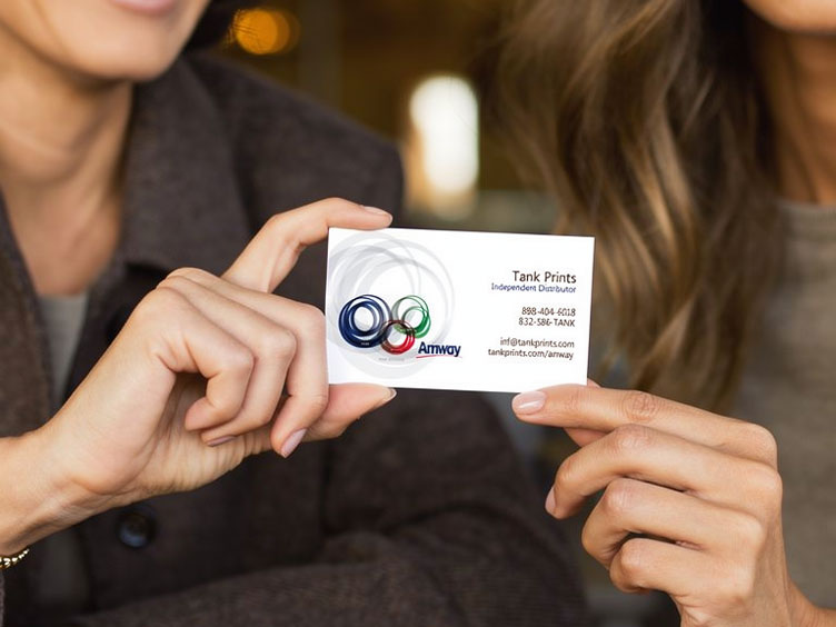 Two people holding a white business card featuring the Amway logo and contact details, with a blurred indoor background.