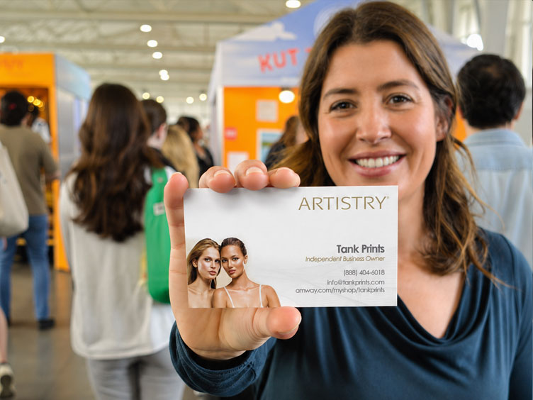 A smiling woman at an event holds up a business card for Artistry Amway Tank Prints, with a blurred crowd and booths in the background.
