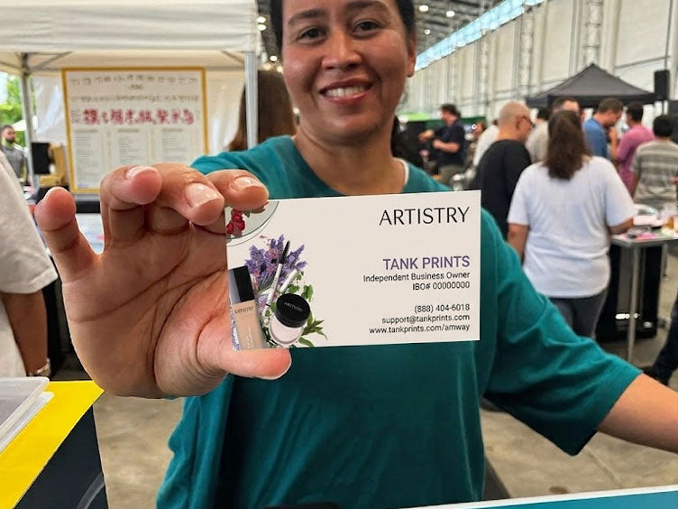 A smiling woman in a teal shirt holds up a business card for Artistry Tank Prints at what appears to be a trade show or event. Amway Cards